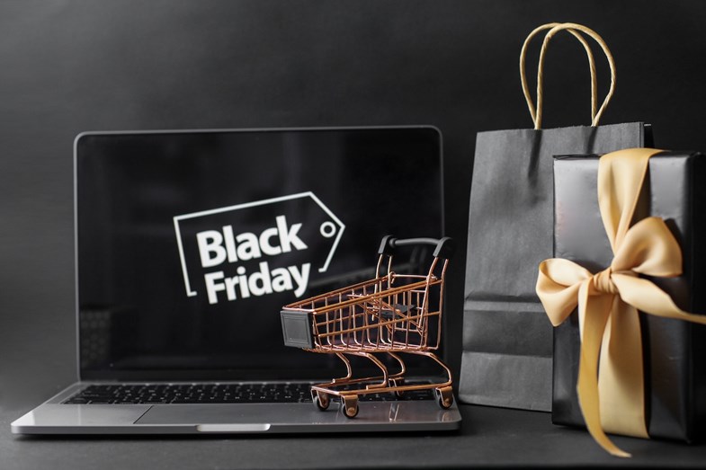 Computador portátil com wallpaper alusivo à ‘Black Friday’, em que um carrinho de compras em miniatura está em cima do teclado e dois sacos de prendas estão ao lado do ecrã
