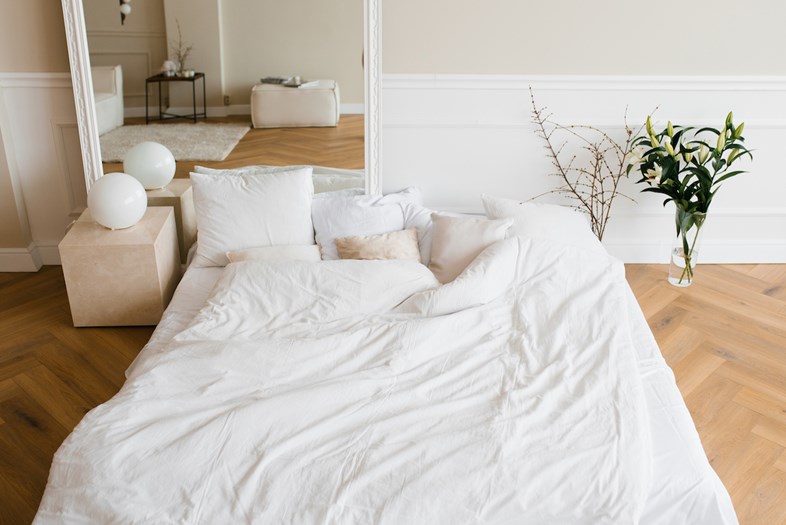 Uma cama de casal com edredão branco num quarto com um espelho e flores no chão.