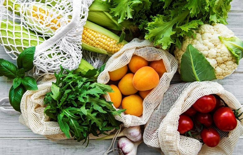 Vários tipos de legumes e frutos saudáveis, separados por sacos sustentáveis, em cima de uma mesa.
