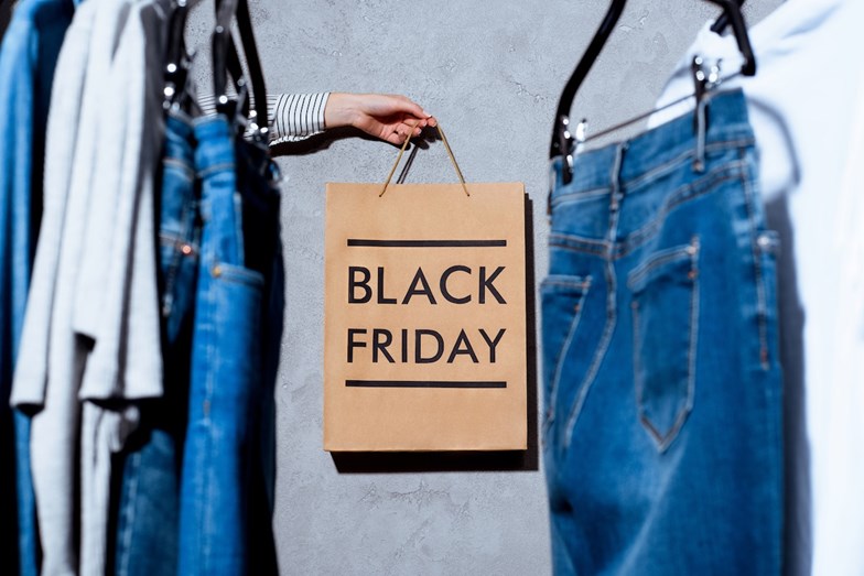 Mão segura um saco de papel com a inscrição “Black Friday” entre roupas penduradas num cabide