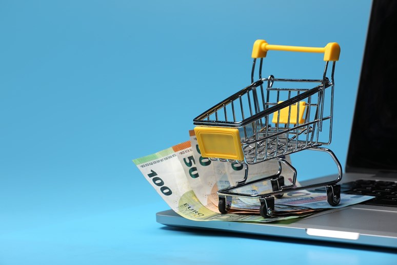 Carrinho de compras em miniatura em cima de um computador portátil e notas de dinheiro.