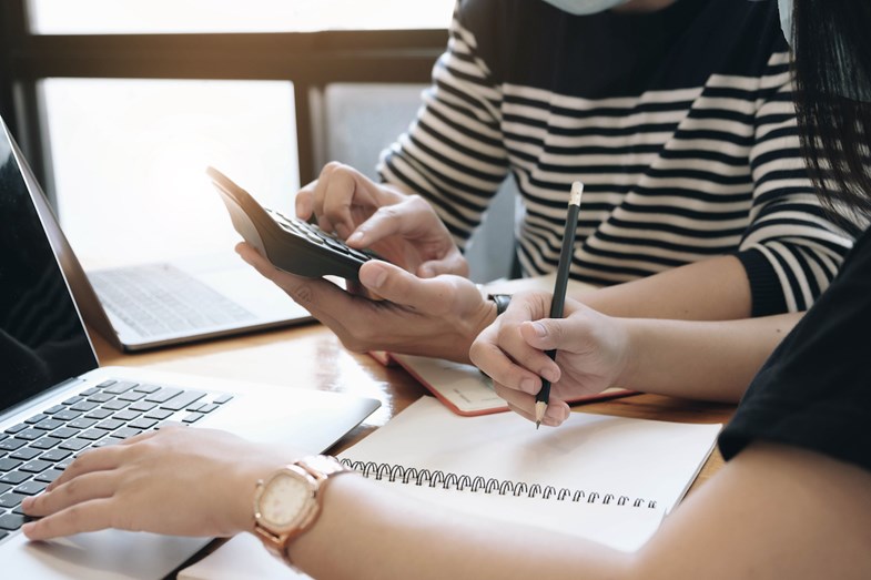 Homem e mulher a fazer contas numa com recurso a uma calculadora enquanto anotam os resultados num bloco de notas e no computador.