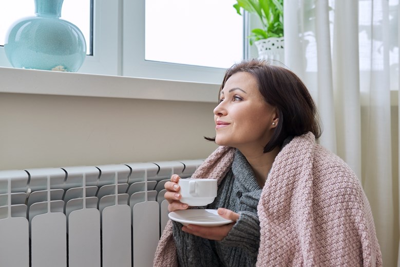 Uma mulher sentada no chão segura uma chávena de café ao pé de um radiador enquanto olha pela janela.