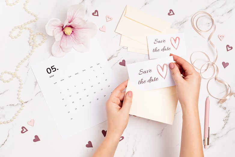 As mãos de uma mulher seguram um cartão ”save the date” sobre uma mesa de mármore, com elementos decorativos de casamento.